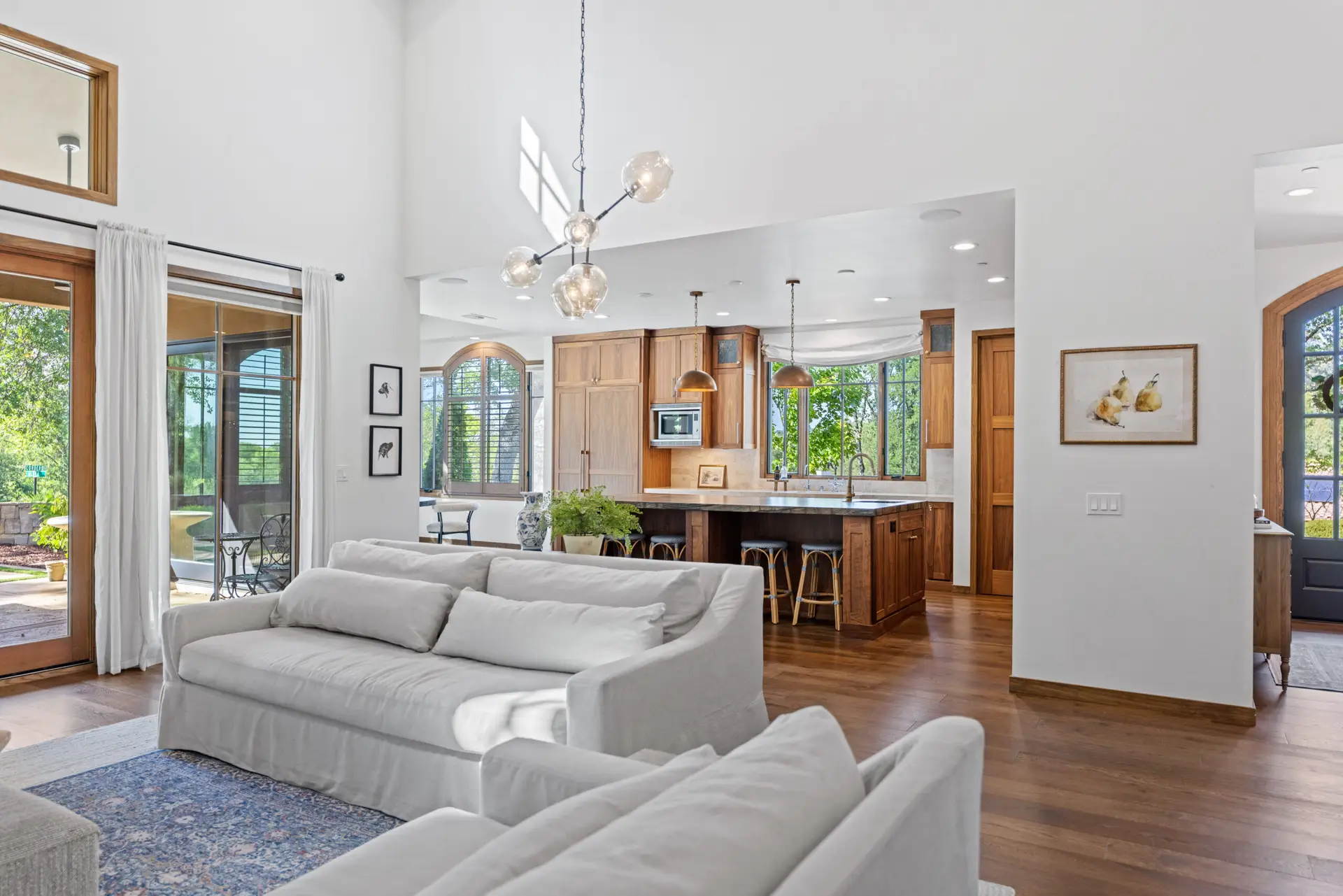 Interior real estate photo of a bright Serrano living room and kitchen with wood cabinets, modern lighting, and large windows.