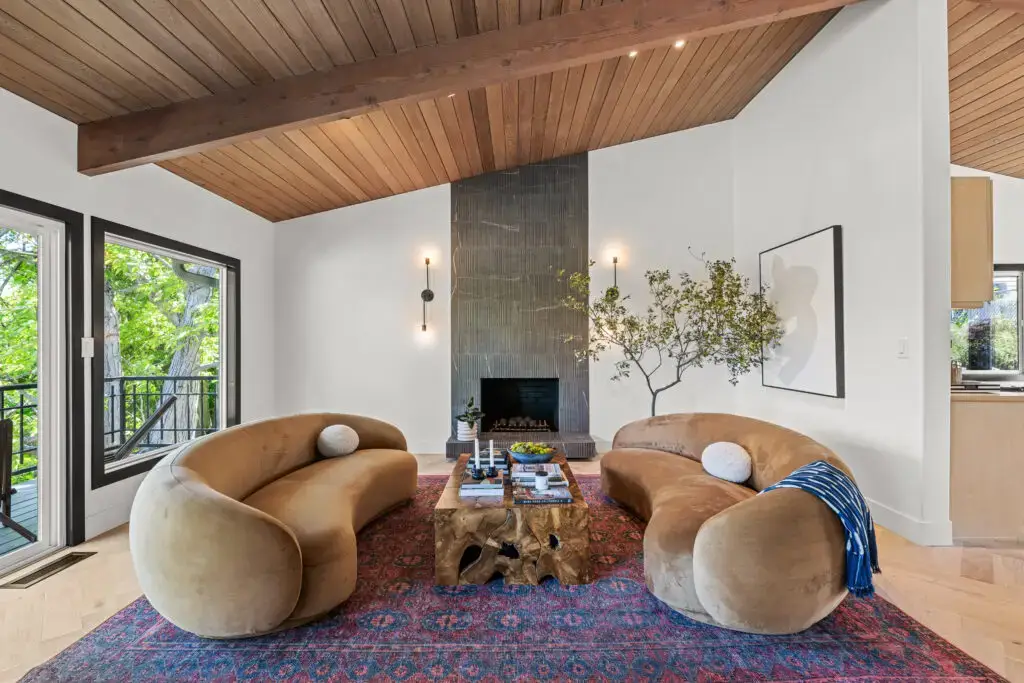 Interior real estate photo of a modern living room in a Carmichael home featuring curved sofas, a tall fireplace, wood ceiling accents, and large windows with natural light.